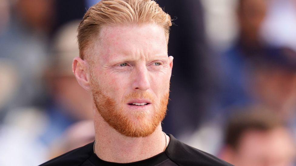 Close up portrait of Ben Stokes wearing a black top. He has red hair and a beard and is looking slightly to the right. Behind him in the distance is a blurred crowd of spectators.