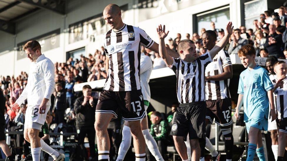 Footballers walk onto a pitch as fans cheer behind them on a sunny day. They are accompanied by mascots. One set of players is wearing black and white striped kits, the other is wearing all white.