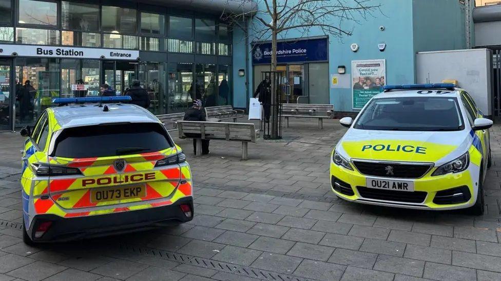 Two neon-striped white police cars parked on a paved pedestrian area outside a bus station, which is a large glass fronted building. Next to it is a small office with a blue sign saying "Bedfordshire Police". There are  wooden benches and a tree between the cars.