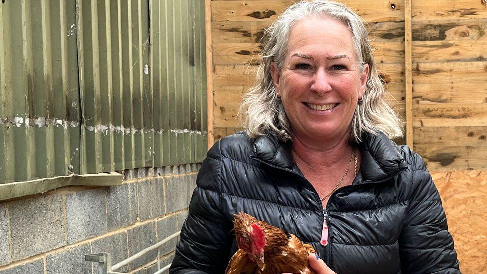 Dawn French in a farm shed/barn, wearing a black puffa jacket and holding a brown hen. She has shoulder-length grey hair and is smiling broadly at the camera. Behind her can be seen a pile of hay.