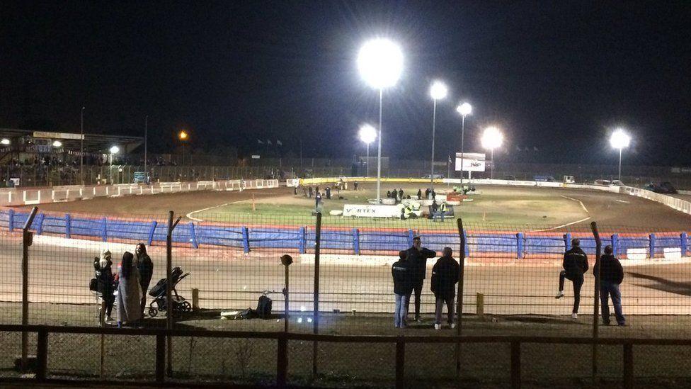 The Arena Essex Raceway. People are standing behind a fence looking towards an oval dirt track. Several bright floodlights are on. Speedway riders and bikes can be seen on and near the track.