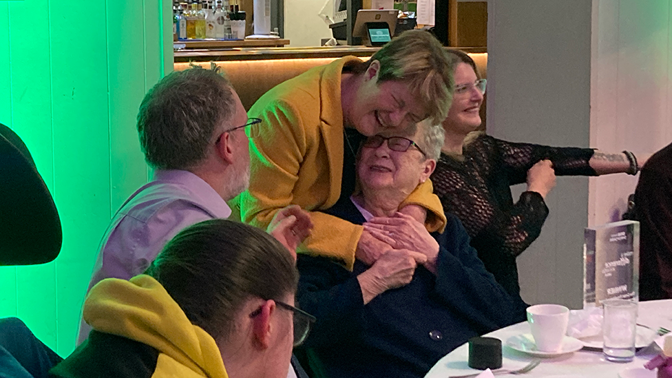 Pat Snow in a yellow jacket hugs Pam after receiving her award. pam is sitting at the table, surrounded by other guests