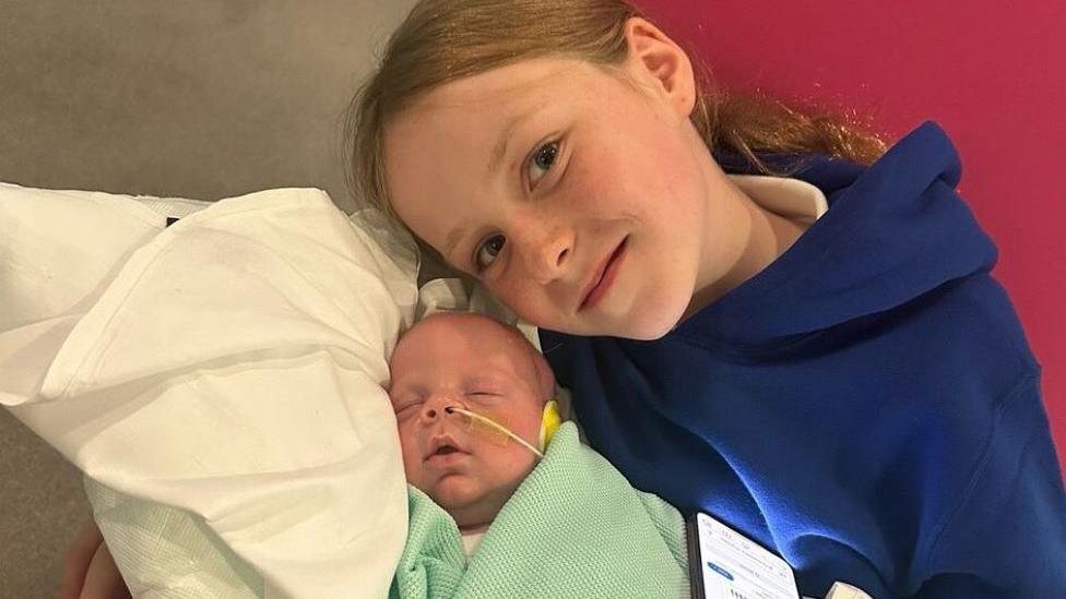 Kobi is lying on a pillow in a green blanket. He has a feeding tube in his nose. His sister is wearing a blue hooded top. She is smiling at the camera and has her face near her baby brother. She has long brown hair.