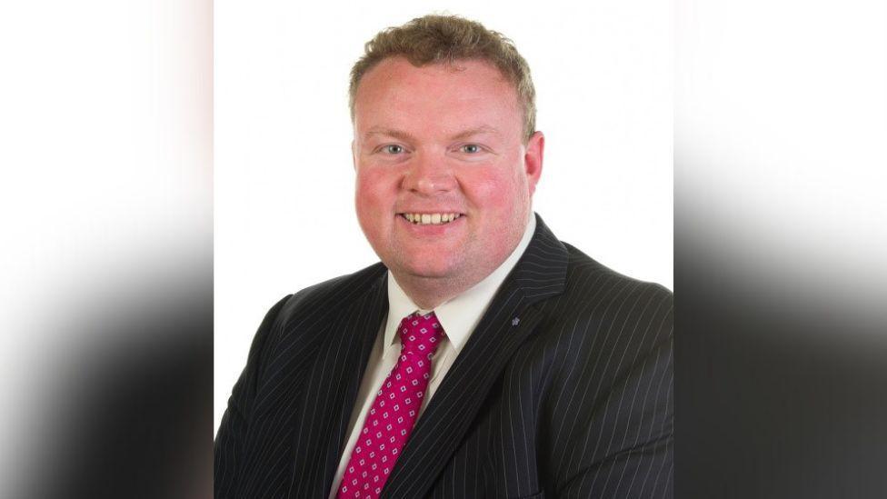 Keith Wysner smiling wearing a pinstripe blazer, a white shirt and a red patterned tie.