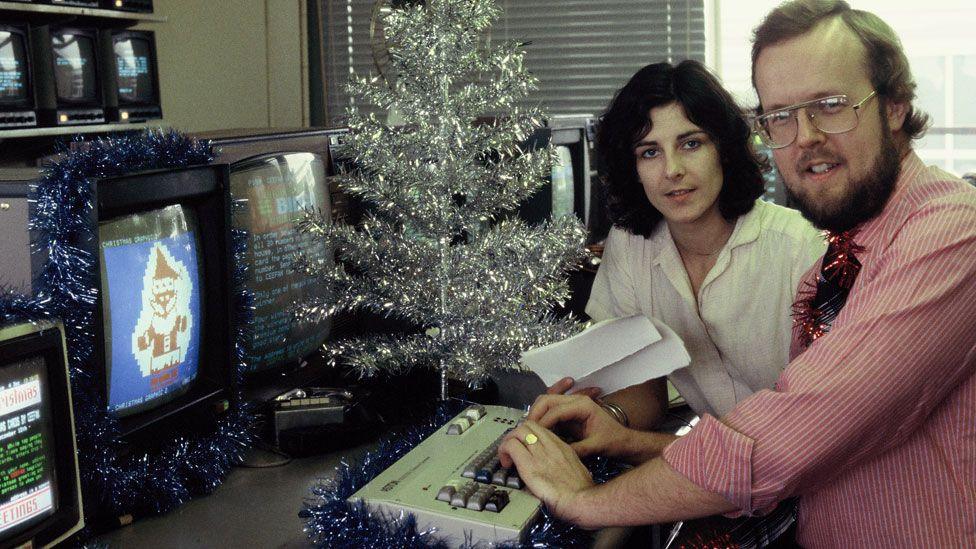 Liz Holdaway with dark shoulder-length hair and Ian Morton-Smith with a beard and glasses, at a desk with a Ceefax keyboard in front, several monitors, one showing a Ceefax Father Christmas design, and a silver Christmas tree on the desk