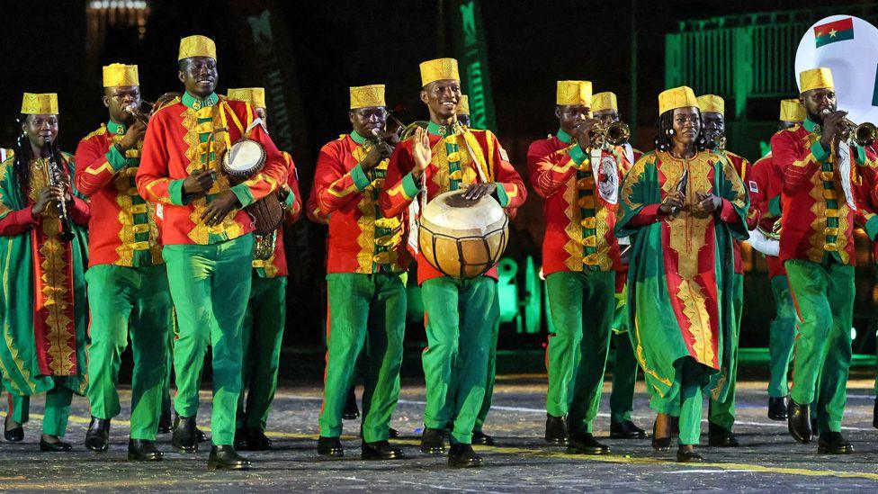 An orchestra from Burkina Faso - its members dressed in green, red and yellow with yellow hats -march and perform in Moscow's Red Square. They are playing a mixture of wind, woodwind and percussion instruments.