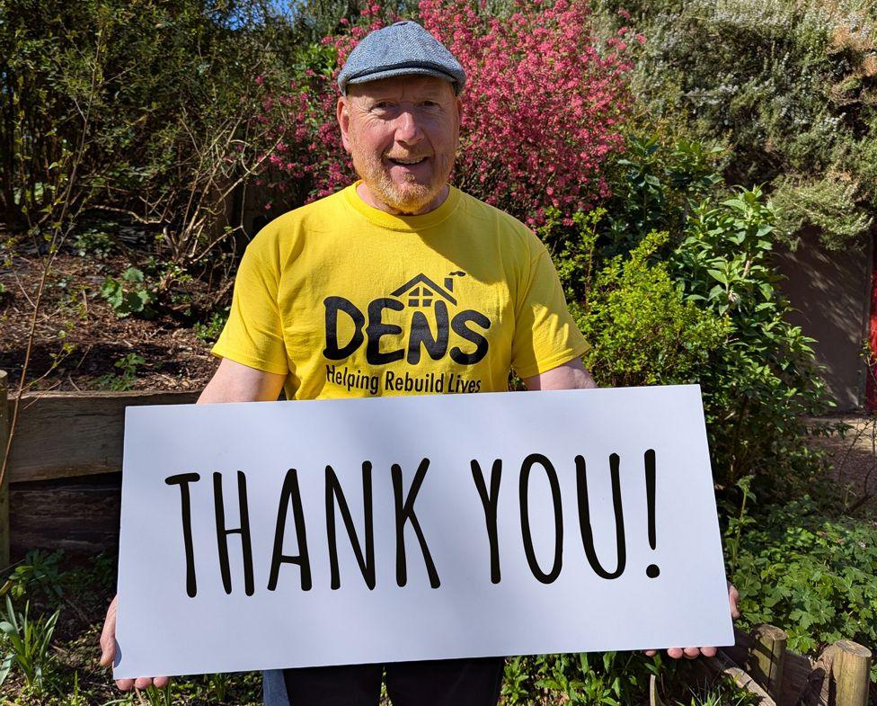 A man wearing a yellow T-shirt and a cap. He is holding a large sign saying "THANK YOU!"