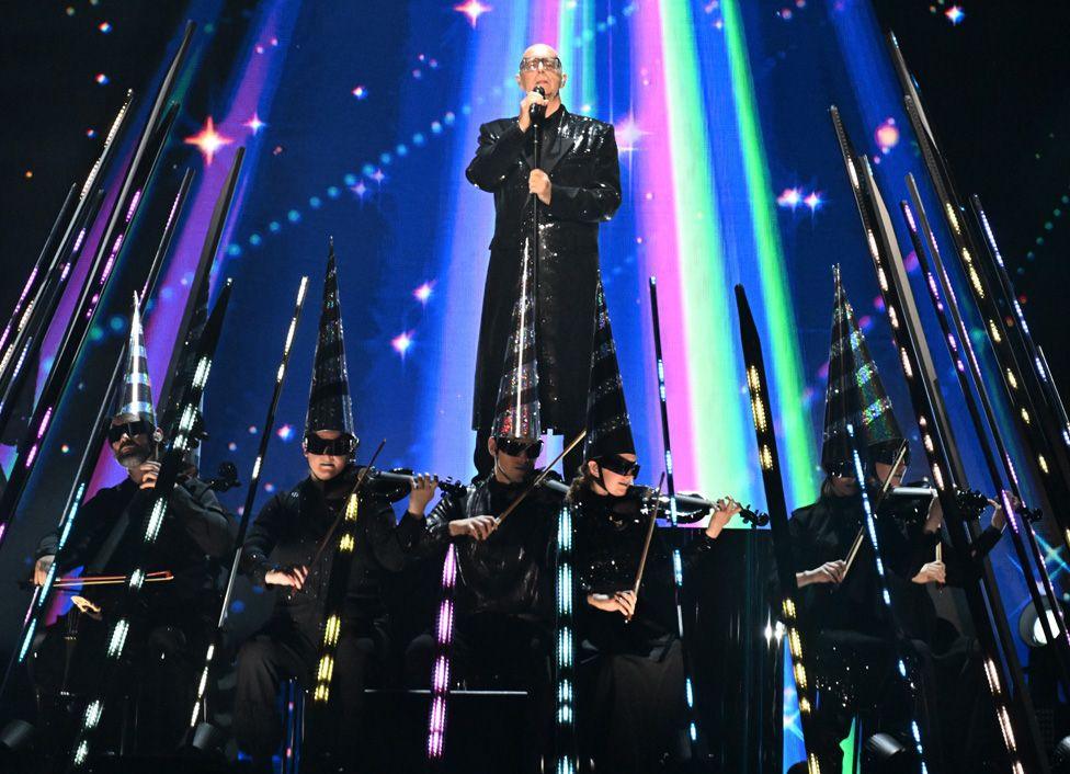 Neil Tennant standing on a platform in the middle of rays of multi-coloured lights, above a row of violinists wearing tall pointy black hats