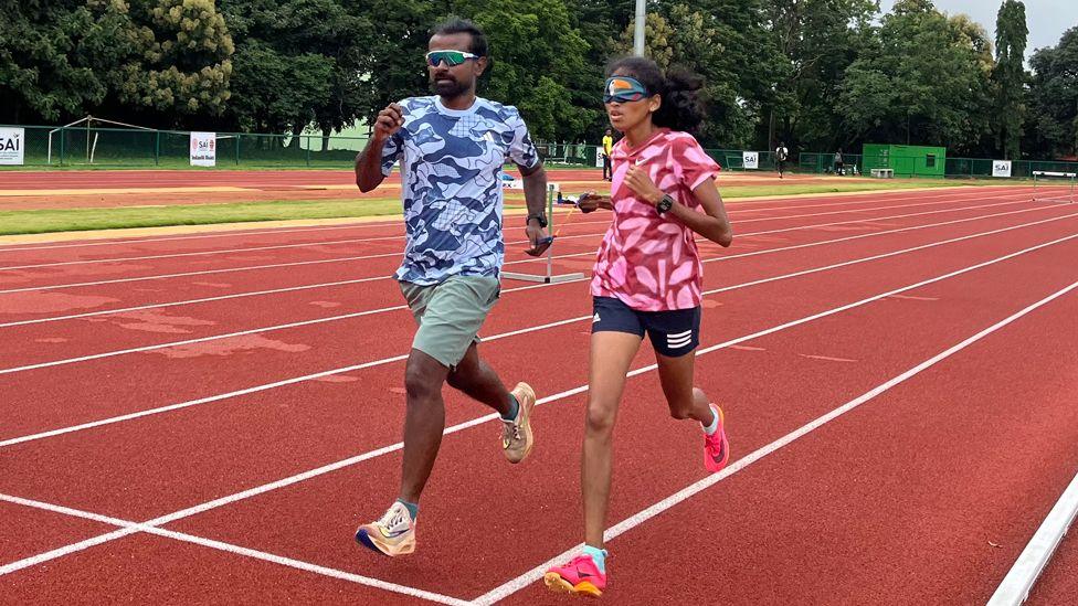 Rahul and Rakshitha running side by side on a track, their arms joined by a tether. He is wearing sun glasses and she is wearing a mask over her eyes. Both are wearing shorts. 