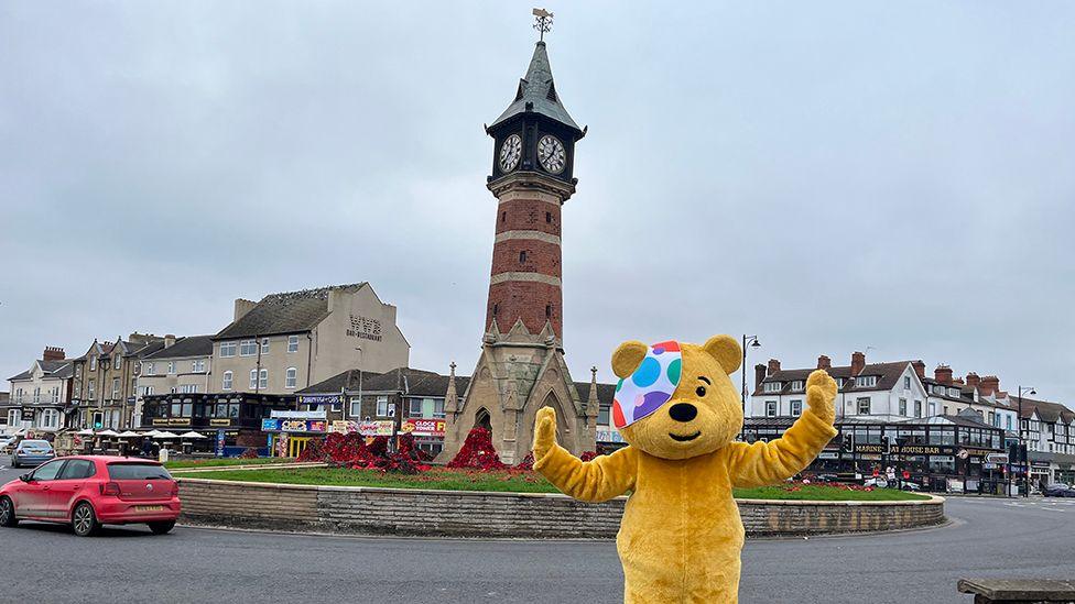 Pudsey posing in front of the clock tower in Skegness which is a brick column with a four-faced clock surrounded by poppies on a roundabout