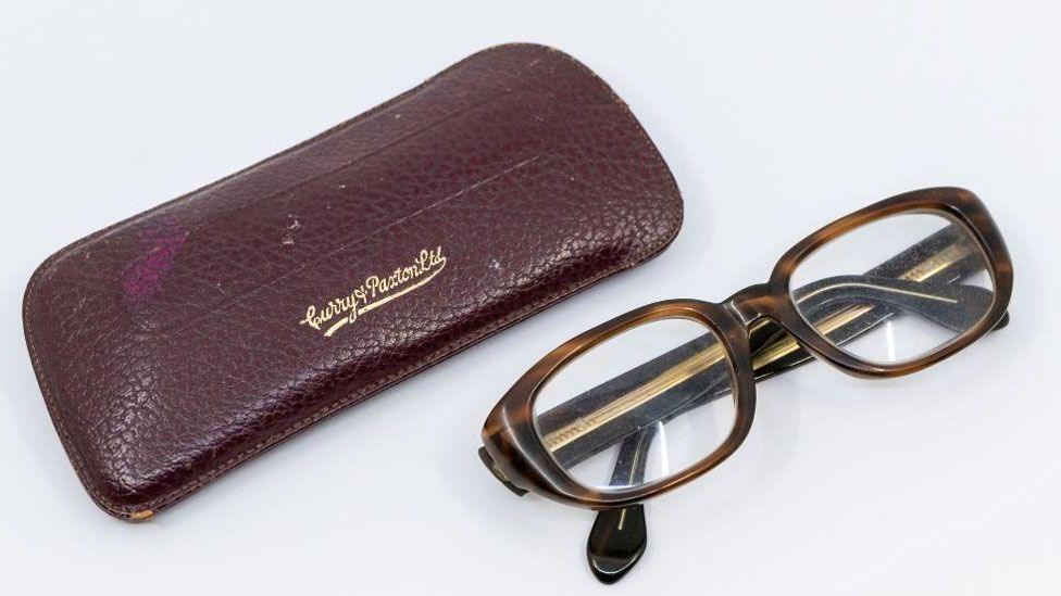 A maroon leather glasses case on the left and to the right is a pair of horn rimmed heavy glasses, folded up on their arms