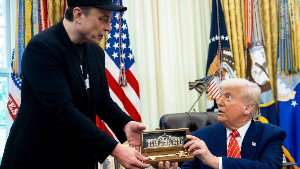 Donald Trump gifts Elon Musk a large key inside the Oval Office. Trump wears a blue suit and red tie, while Musk wears a black suit jacket and pants with a black 'Doge' hat.