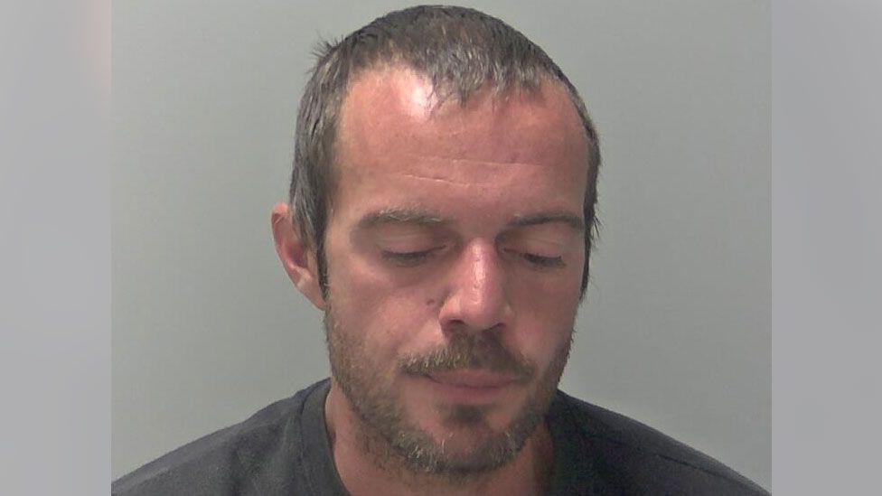 A man, Daniel Gibbs, looks down at the ground in a police mugshot. He has light brown, short hair and light brown stubble. He is wearing a dark-coloured top.