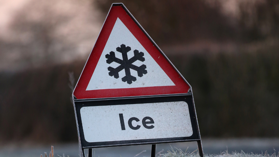 A road sign warning of ice,  The sides of the road are covering in snow. 