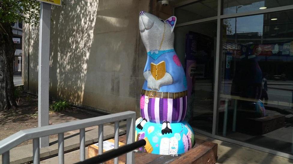 A sculpture with lots of purple and blue colours and also holding what appears to be a book is also displayed next to the front entrance of a building