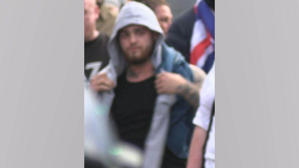 A blurred image of a man in a hooded top, with a brown beard and dark hair, who is wanted by police. He is wearing a black top and grey hoodie, and has a sleeve tattoo down his left arm.