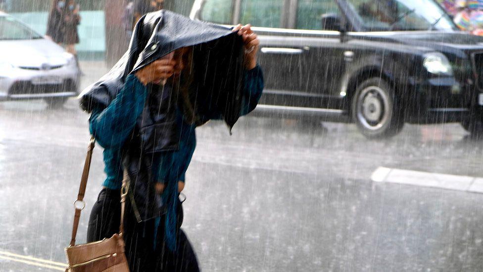 A woman holds her jacket over her head as rain pours down in London