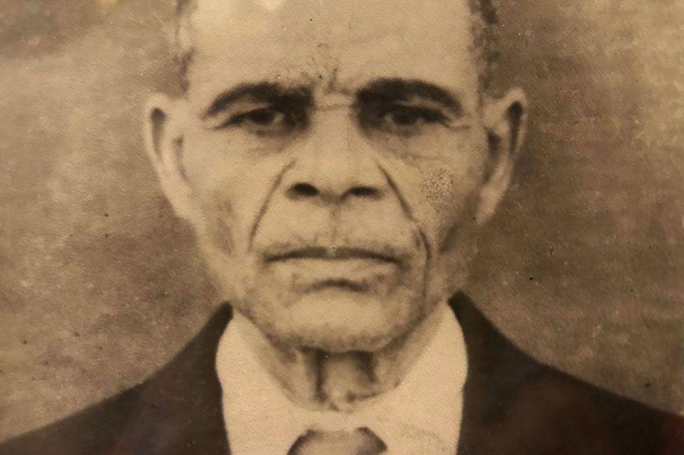 A old sepia Carmen Souza's grandfather as a older man. He is staring straight at the camera, and is wearing a suit and tie