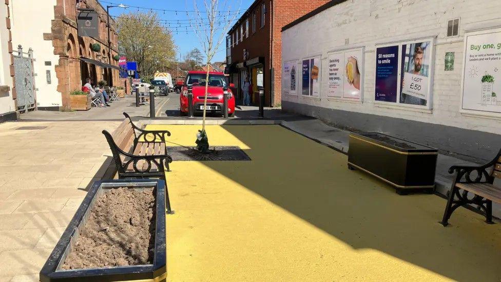 An area with benches and a large yellow section of ground. There is a car in the background and a coffee shop. It is a bright day and there is some shade from a high wall.