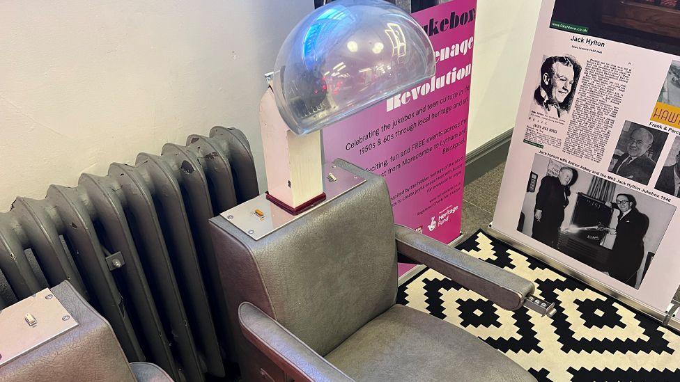 An old-fashioned helmet-style hairdryer attached to a grey barber chair. To the left is a large grey radiator and to the right are posters advertising the art installation