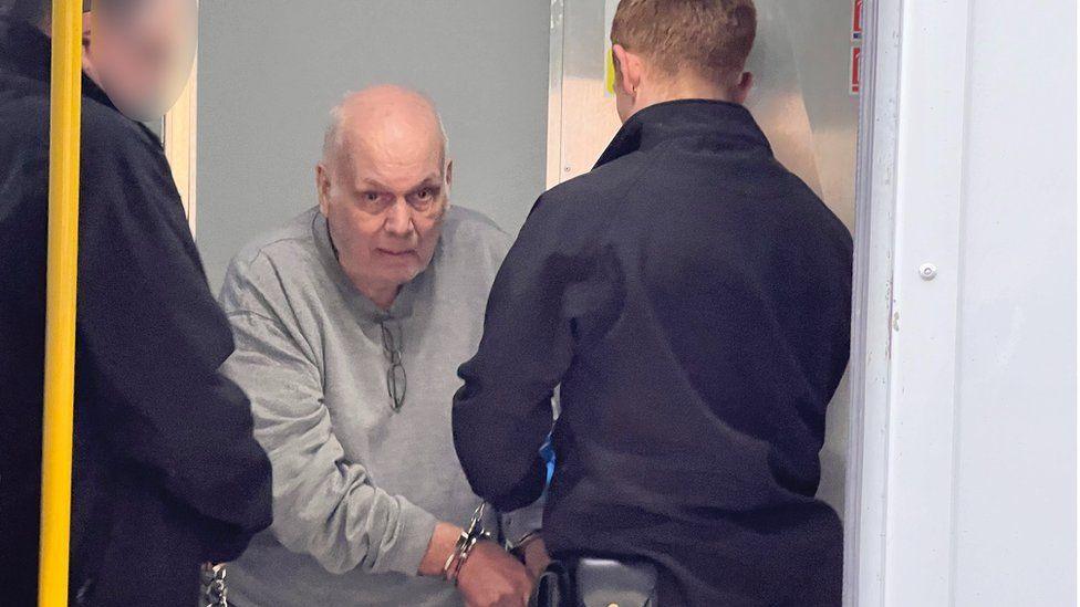 A man in a grey jumper with handcuffs around his wrists looks towards the camera. Two police/prison staff are in the image with their backs to the camera