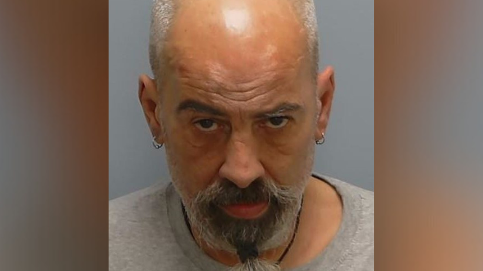 Kenneth Walker pictured in a mugshot. His head is looking down but his eyes are fixed on the camera. He has a bald head with short grey hair at the sides, ringed earrings in both ears, and a thick black and grey goatee beard tied at the bottom.