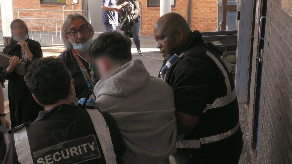 A television still, showing members of hospital staff, including Leston Scafe, holding a young man in a grey hoodie by the arms