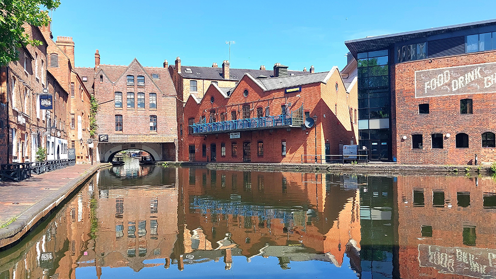 The canal in Birmingham on a sunny day. Two- three- and four-storey buildings made of red brick flank the canal on either side as well as in the distance, built over a short tunnel to allow boats to pass through