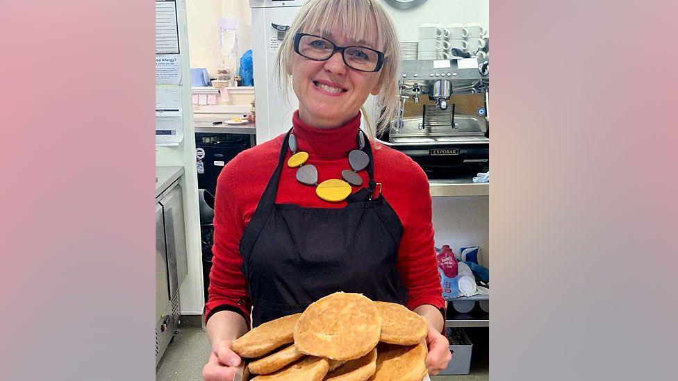 Iryna Porfilova holding a tray of butteries. She is wearing a red turtle neck jumper, navy apron, and a yellow and grey necklace. She wears glasses and is smiling in the photograph. Behind her is a coffee machine and a kitchen.