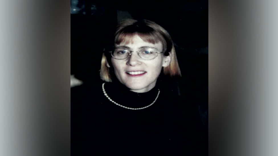 Headshot of woman smiling at the camera. She is wearing a black turtleneck, glasses and a necklace.