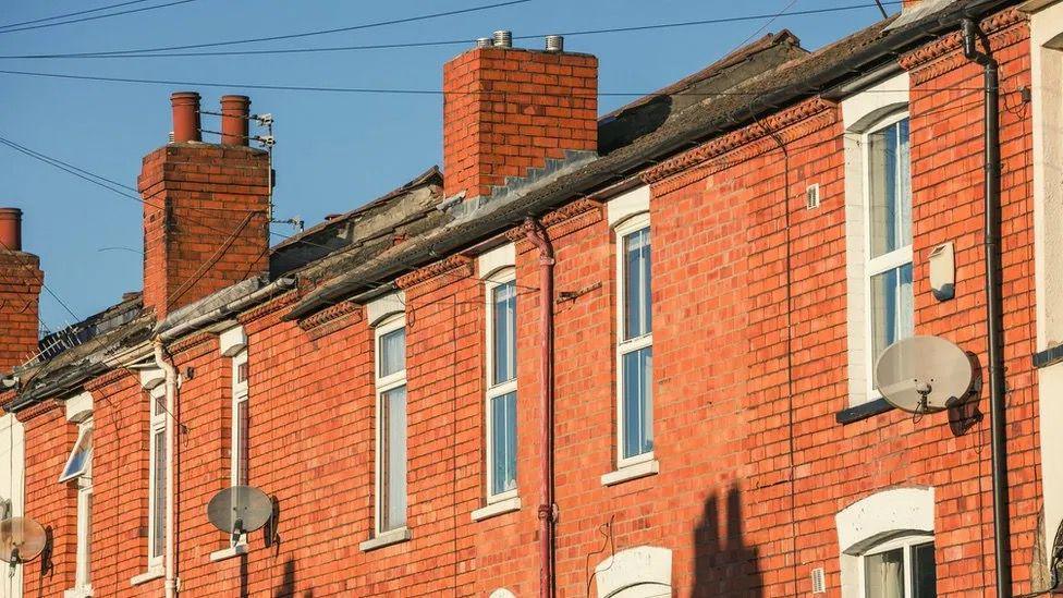 The outside of red bricked terraced homes with white windows, chimneys and satellite dishes. A blue sky can be seen at the back of the homes.