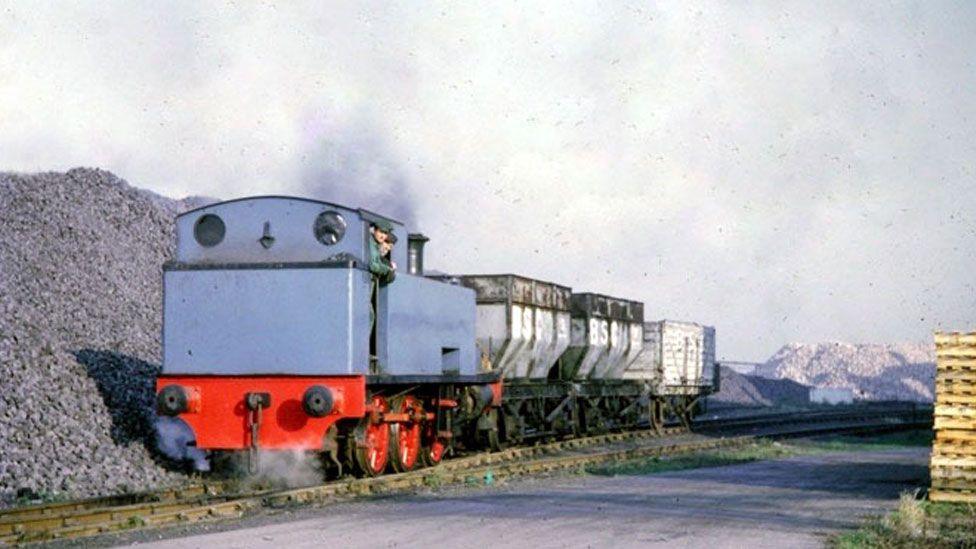 A grainy colour photo showing a Hudswell Clarke locomotive running on lines. It has a light blue body and red undercarriage. Two men wearing caps can be seen looking out of its side door. Behind it are three white railway trucks. To its right is a large grey pile of something - possibly cole.  