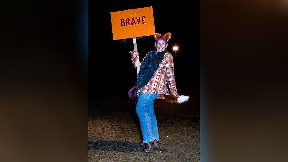 A woman wearing an orange checked shirt, blue jeans, a pair of fox ears and a fox tail. She's striking a pose, smiling, and holding up a sign saying "brave".