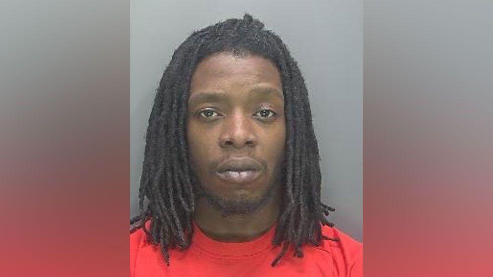 Police custody image of Omar Harris. He has shoulder-length dreadlocks and is wearing a red T-shirt.