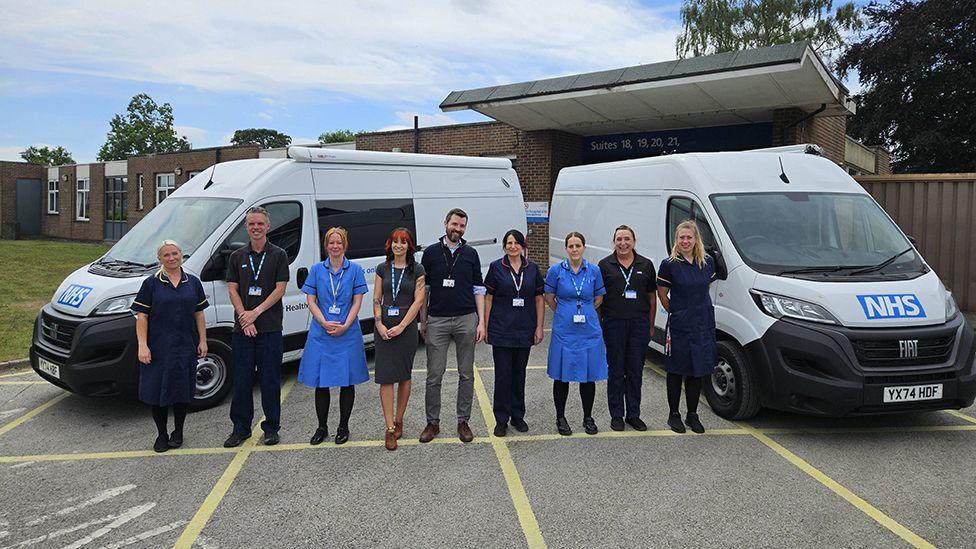 NHS staff – some wearing medical uniforms – lined up between the white vans, which have NHS logos on the front.
