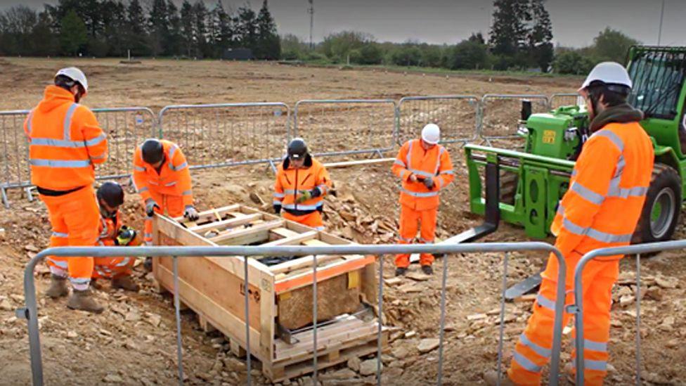 Team of workmen wearing red high-visibility suits standing around the grave with a metal fencing around them