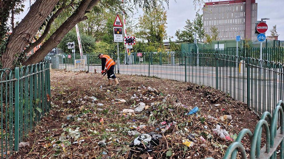 A worker in a high-visibility jacket cleans up rubbish and plant waste covering a curved piece of land at the side of a road with green metal railings bordering the road behind.