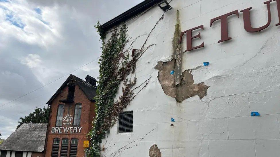 In the foreground is the front wall of the Three Tuns Inn - the white plaster is cracked and peeling off, and the sign is falling apart, so it just reads 'E TUN' in dark red lettering. Ivy is growing up part of the wall. In the background, the Three Tuns Brewery can be seen with its name painted on the red brick building in white lettering.