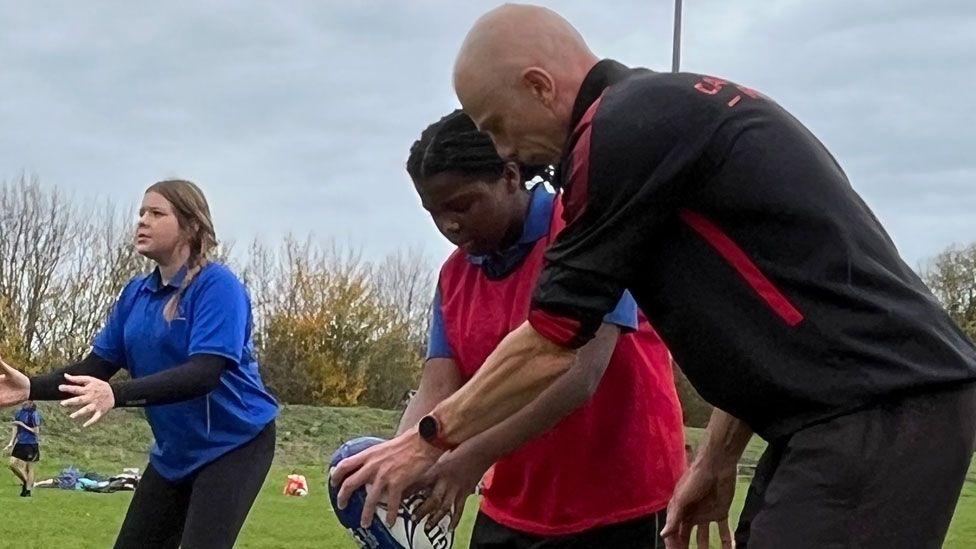 Three people on a rugby pitch. On the right is a man, sideways on, in a black kit standing beside a girl in a red bib over a blue short-sleeved shirt. They are both bending over and looking towards a rugby ball, held by the girl. The man also has a hand on the ball. On the left is a girl in a blue short-sleeved shirt holding out her hands ready to catch something.