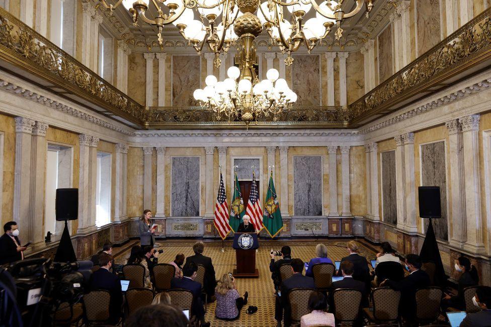 A large gilded room with then Treasury Secretary Janet Yellen addressing an audience. There are neo-classical pillars lining the walls and marble inlays and a gold mezzanine balcony halfway up the cavernous walls.