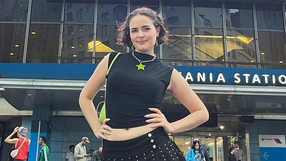 Kelley Heyer, a woman, outside Madison Square Gardens, wearing a black top with a lime green star shaped necklace. Her hands are on her hips, with other concert-goers in the background.