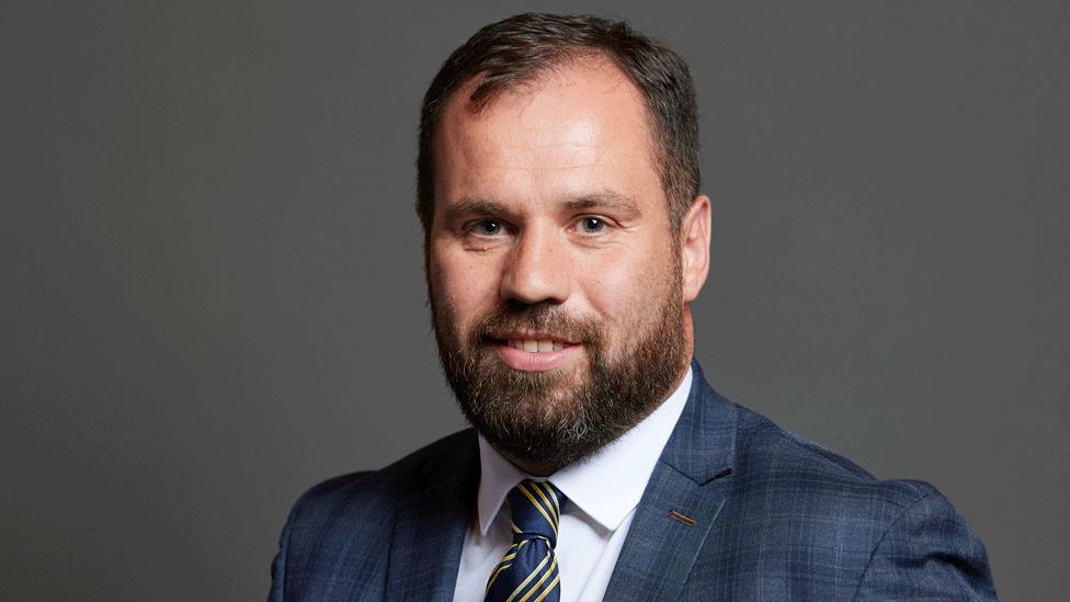 Sean Woodcock's parliamentary photo portrait. He has short brown hair and a brown beard. He wears a blue suit, blue and gold striped tie, and white shirt.