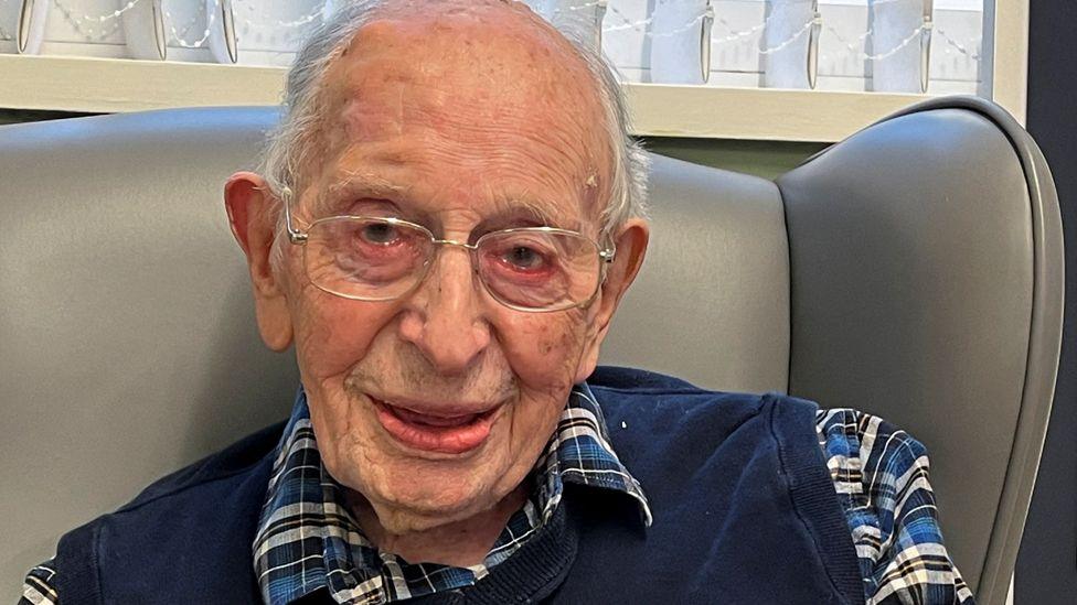 John Tinniswood, pictured with a blue checked shirt and blue tank top, smiling at the camera at his care home in Southport