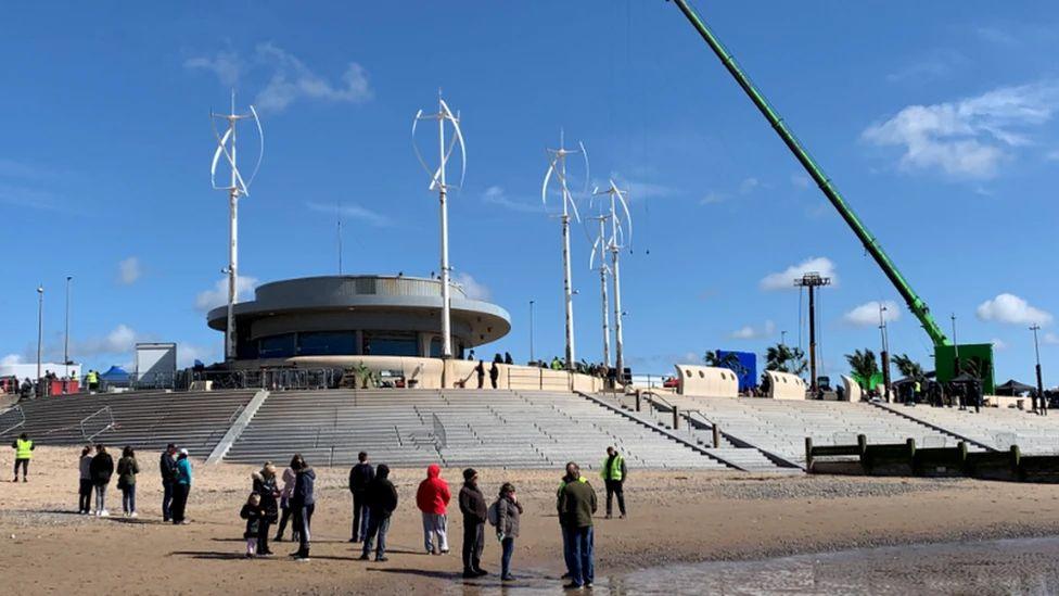 The FBKafé on the seafront, a round single storey cafe with steps down to the sand. It's an unusual looking building and is similar to a spaceship. People are standing round on the sand 