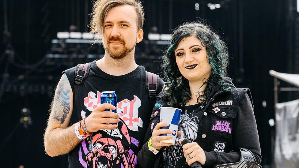 A man and a woman stand in front of an empty festival stage, each holding a drink and smiling. Both are dressed in alternative styles - the man has an asymmetrical haircut and wears a black sleevless top with a band's logo on it. The woman wears a black denim jacket with patches for Volbeat, Black Sabbath and System of a Down