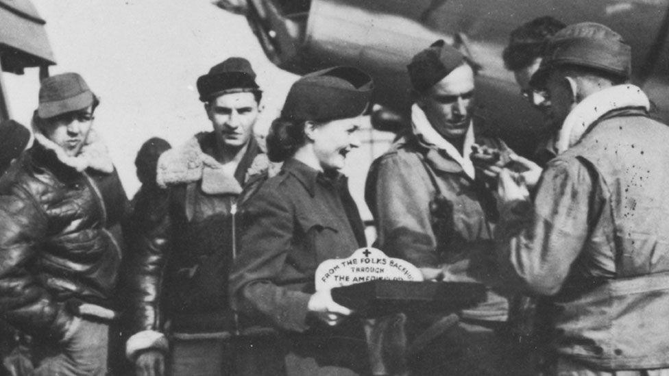 An American Red Cross woman serving five US airmen during World War Two somewhere in the the UK. She is in the middle and sideways on, smiling towards three of the men. She has a tray in front of her. Behind the group can be just seen a B-17 Flying Fortress bomber. 