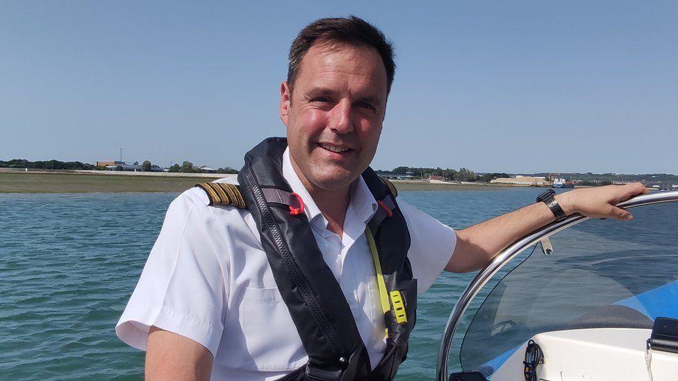 Harbourmaster Billy Johnson smiles on board a small boat at sea. His uniform includes a white shirt with epaulettes and he wears a lifejacket.