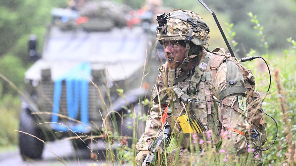 A soldier crouching by the side of the road  in camouflage uniform. Behind him there is an armoured vehicle, slightly out of focus