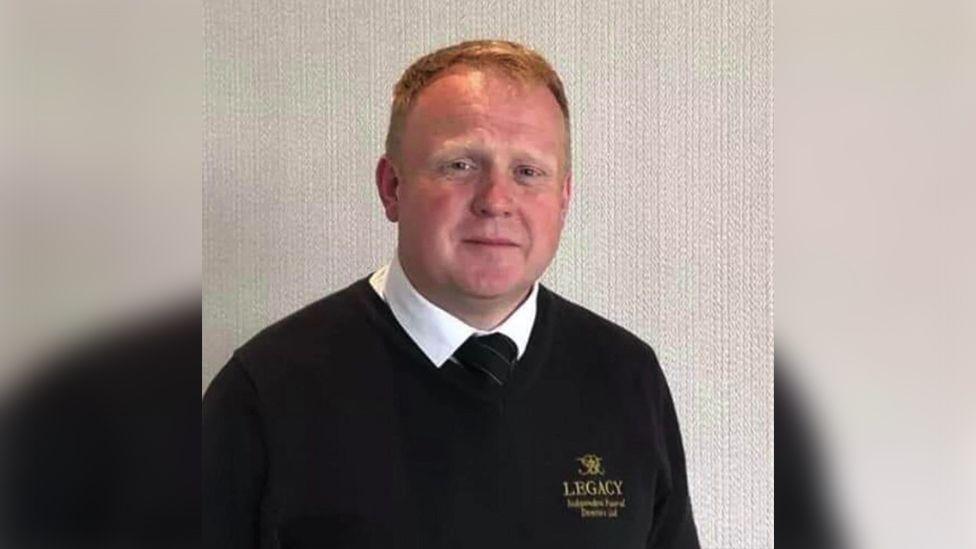 Robert Bush has short ginger hair. He wears a white shirt, black tie and a black jumper.