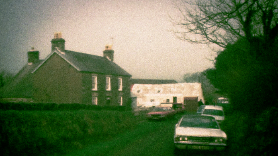 Archive image from 1976 of a grey farmhouse on a lane, with a white barn next to it and lots of old police cars parked outside